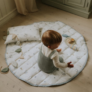 Child sitting on a round quilted mat with toys on a wooden floor.