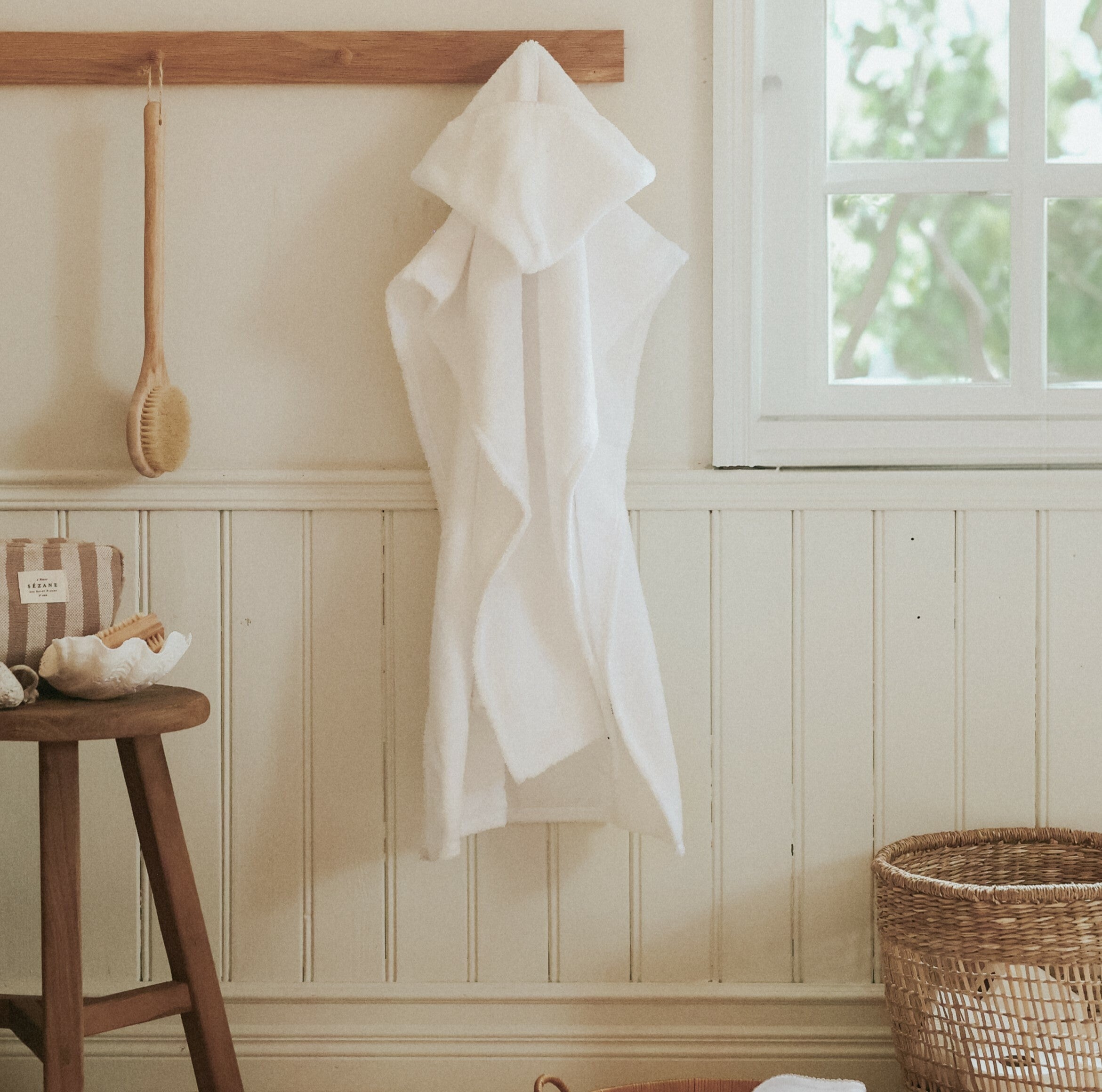 Toddler Poncho hanging up on the wall in a bathroom. A wooden stool with bath accessories to the left and a rattan storage basket with towels to the right.