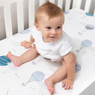 Crib Sheet in "Touch The Sky" print in the color blue featured in a crib with a baby sitting on top smiling