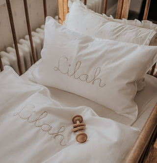 White Bedding and white pillow with a personalized monogram on both blanket and pillow. Wooden rattle laying on top of bedding