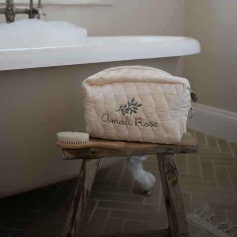 Toiletry Pouch in Ivory sitting on top of wooden stool in the bathroom next to a bathtub with bubbles in it. Wooden hair brush sitting next to Toiletry Pouch