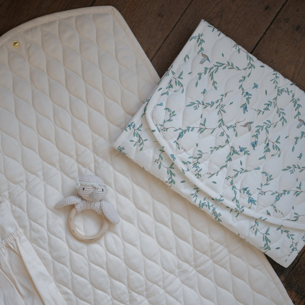 Portable Changing Pad in Ivory opened up laying flat on the floor. Inside is in Ivory with pocket detail and a folded changing pad in Secret Garden laying on top with a knit gooseling rattle.