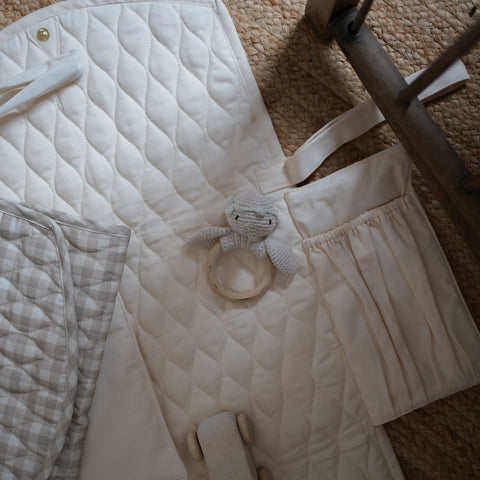 Portable Changing Pad in Ivory opened up laying flat on the floor. A folded changing pad in Beige Gingham laying on top with a knit gooseling rattle