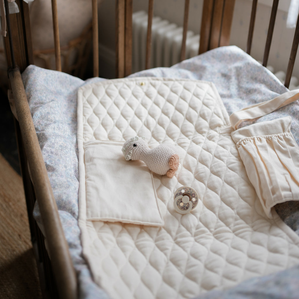 Portable Changing Pad in Ivory opened laying in a crib showing two pockets with a knit gooesling stuffy and pacifier laying on top.
