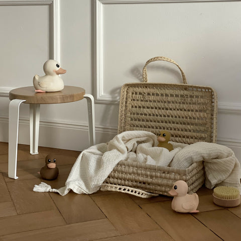 Rubber Gooseling Bath Toy in Marshmallow sitting on a wooden stool. A rattan open suitcase with cream blanket and three additional ducks playing inside 