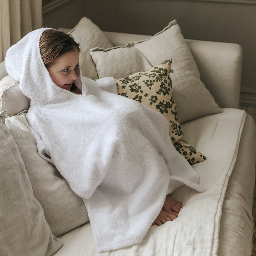 Toddler Poncho in Beige. Child sitting on couch with Toddler Poncho on with hood on her head.