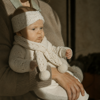 A baby wearing a cream-colored knit headband, matching scarf with pom-poms, and a cream knit outfit, held by an adult in a cozy brown sweater in warm, direct sunlight.