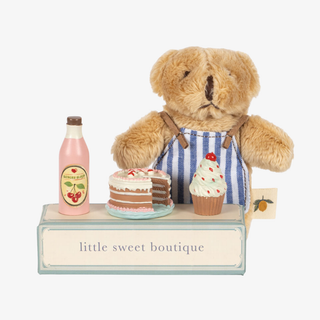 Small tan teddy bear standing behind toy deserts on a cardboard table that says "little sweet boutique". 