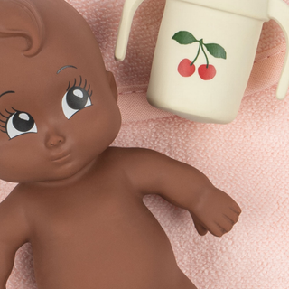 Brown-skinned baby doll with large eyes next to a white cup with red cherries on a pink textured background