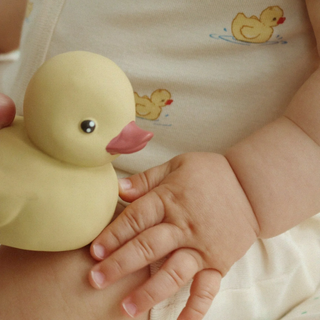 Yellow rubber duck held by a child's hand with a baby's outfit in the background