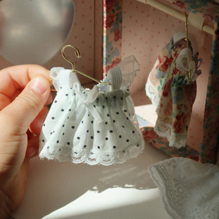 White lace dress with black polka dots on a gold hanger held by a hand in a dollhouse setting.