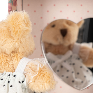 Teddy bear wearing a polka dot dress looking at its reflection in a mirror.
