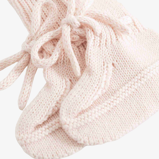 A close-up of a pair of pale blush pink knit booties with matching laces, showing the detailed cable-knit texture