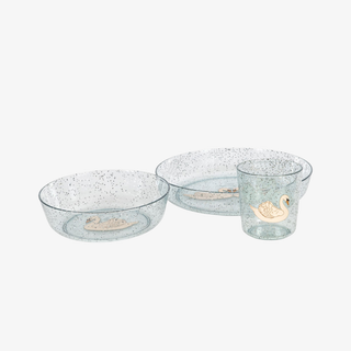 A coordinated children's tableware set on a light gray background, including two round plates and a small tumbler. The pieces are translucent pale blue-gray with a sparkle finish and each features a central swan illustration in soft peach and beige tones.