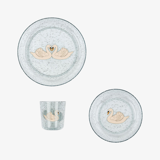 A coordinated children's tableware set on a light gray background, including two round plates and a small tumbler. The pieces are translucent pale blue-gray with a sparkle finish and each features a central swan illustration in soft peach and beige tones.