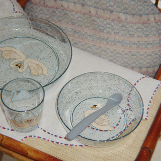 A set of clear speckled children's tableware, including a large bowl, a smaller bowl, a drinking glass, and a spoon, arranged on a cream tablecloth with a patterned cushion in the background.