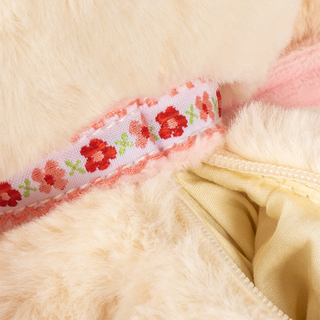 A close up of a cream-colored plush dog purse with a pink patterned strap handle, shown against a plain white background.