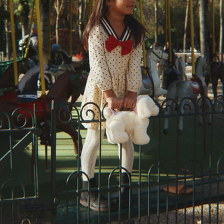 A young girl wearing a cream polka-dot sailor frock dress with a red bow and white tights stands in a park next to a carousel, holding a white plush dog purse.