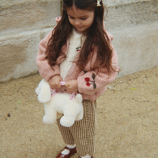 A young girl with long brown hair stands outdoors against a stone wall, wearing a pink fuzzy cardigan with cherry embroidery, brown-and-white check pants, and holding a white plush dog purse.