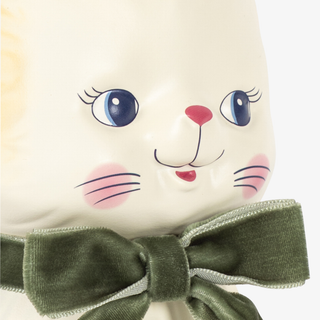 Close-up of a white cat money bank with large blue eyes, pink cheeks, a red nose, and an olive green bow around the neck, shown against a plain light background.