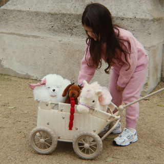 A young girl in a pink tracksuit pushes a small cream-colored toy wagon outdoors. Inside the wagon are three plush stuffed animals: a white dog, a brown dog with a red collar, and a white sheep.