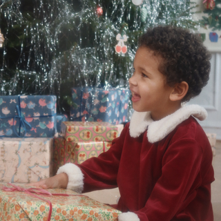 Child in a Santa Claus outfit opening a gift in front of a decorated Christmas tree.