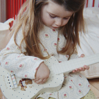A young girl with long light brown hair sits indoors holding and playing a small white guitar with black polka dots. She is wearing a cream-colored long-sleeve pajama top with a small pink floral pattern.