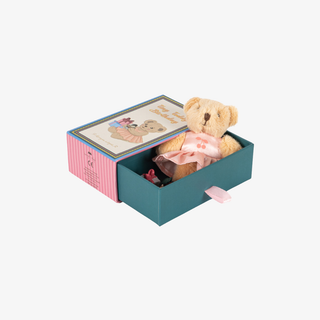 A teddy bear gift set featuring a small plush teddy bear in a pink bow inside an open teal drawer box with a pink striped side panel and a decorative illustrated lid.