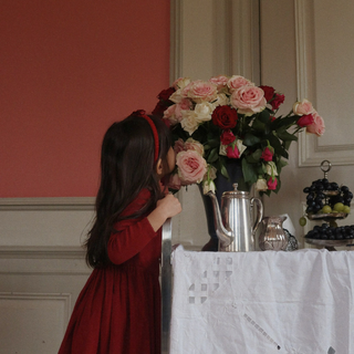 Child in a red dress standing next to a table with flowers and a teapot.