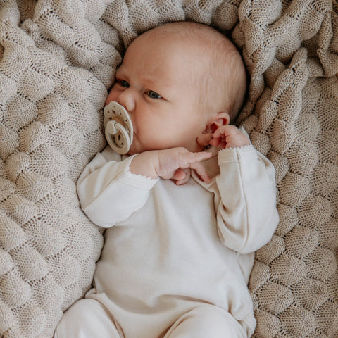 Baby lying on a textured blanket wearing a white outfit and holding a pacifier.