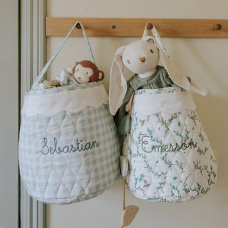 Two hanging storage bags with embroidered names, one with a monkey toy and the other with a rabbit toy, on a wooden hook against a white wall.