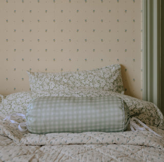 Floral bedding with a picnic gingham green bolster pillow on a bed against a patterned wall.