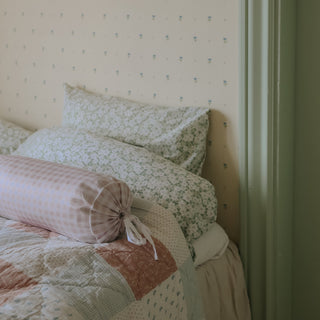 Bed with floral bedding and a picnic gingham pink bolster pillow  against a patterned wall.