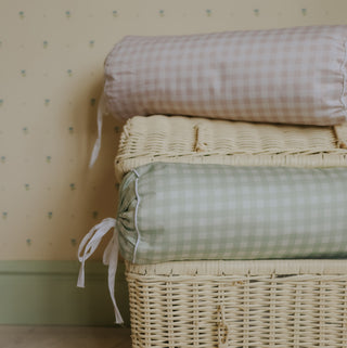 Stack of picnic gingham bolster pillows on a wicker basket against a patterned wall.