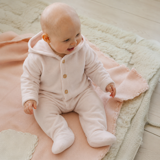 A baby wearing a pale pink footed hooded romper sits on a soft pink blanket over a cream textured rug on a light wood floor.