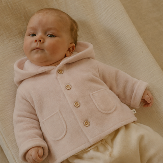 A baby lying on a textured cream fabric surface, wearing a pale blush pink hooded cardigan with buttons and cream pants.