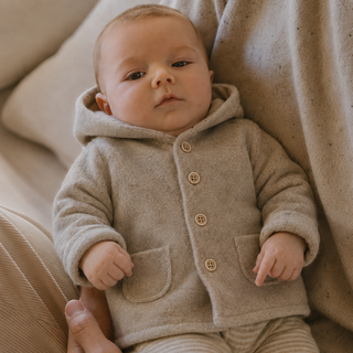 A baby wearing a beige hooded button-front cardigan and matching striped pants, sitting on a person's lap against a soft neutral background.