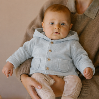 A baby wearing a light blue hooded cardigan and sits on a woman's lap against a warm beige background.