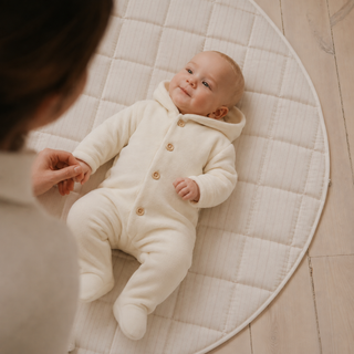 A baby wearing a cream-colored hooded footed onesie lies on a round quilted cream changing mat on a light wood floor, with an adult's hand and arm visible holding the baby's hand.