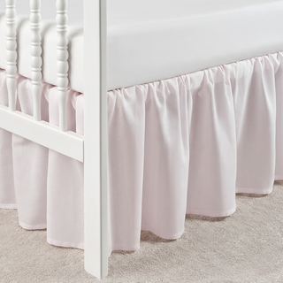 White crib with a light pink ruffled bedskirt on a neutral carpeted floor.