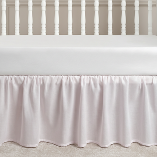 White crib with a light pink crib skirt against a neutral wall. 