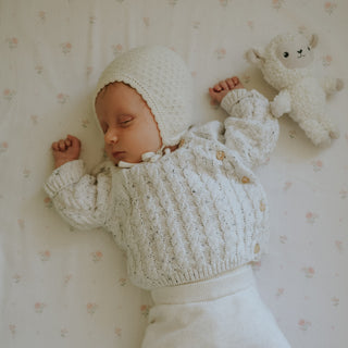 Baby in a white knitted outfit lying on a floral crib sheet with a plush toy.