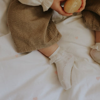 Child's legs wearing brown pants and white socks on a white blanket with pink embroidered hearts.