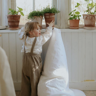 Child in a white shirt and brown overalls standing by a window with potted plants.
