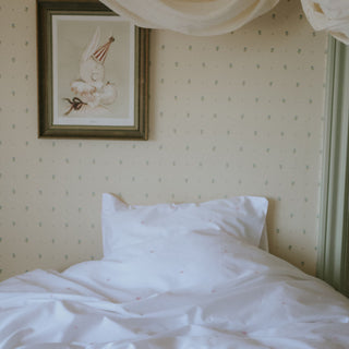 Neatly made bed with white bedding against a wall with decorative wallpaper and a framed picture.
