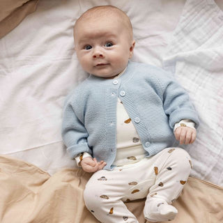 A baby lying on beige and white bedding, wearing a light blue knit cardigan over a cream top and cream footed sleeper with small brown leaf prints.