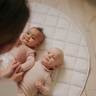 Two babies lying on a quilted mat with a person partially visible.