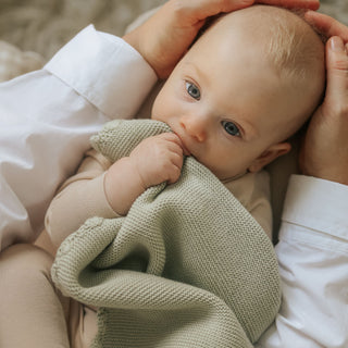 Baby holding a green blanket with a person's hands supporting the baby's head