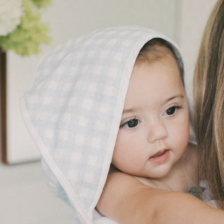 A close-up of a baby wrapped in a light blue and white gingham checkered towel, held by an adult whose arm is visible on the right.