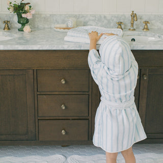 A child wearing a light blue and white vertically striped hooded bathrobe stands in front of a dark wood bathroom vanity with a marble countertop.